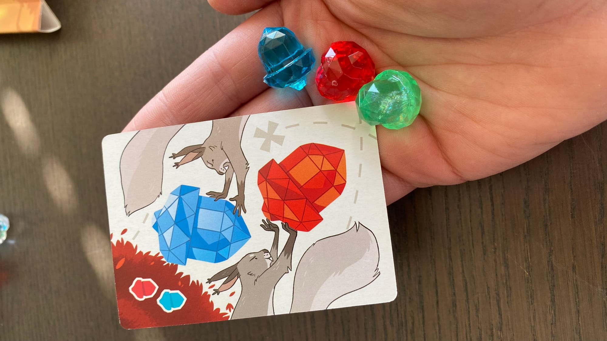 A hand holding a card—depicting one red and one blue acorn—and three acorns colored red, blue, and green